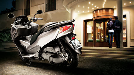 Rear 3-quarter view of left facing Forza 300, parked in front of hotel with rider and pillion going inside.