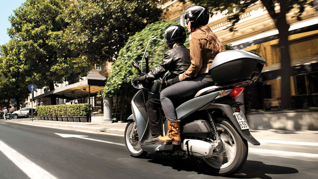 Honda SH125i motorbike rear-threequarter view with rider and pillion. Right-facing (Road location).