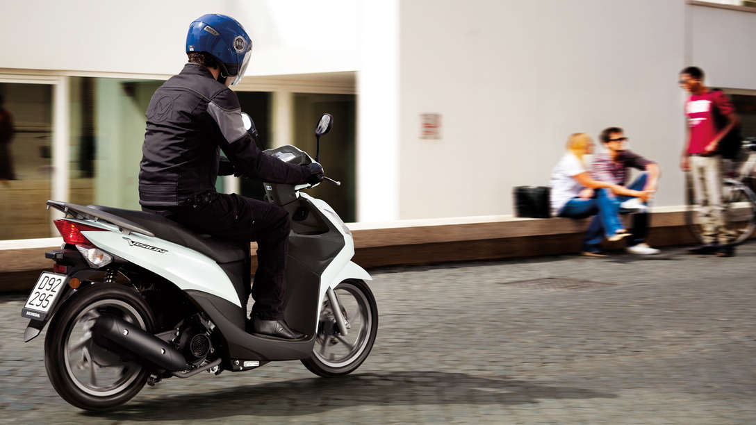 White Honda Vision 50 rear-threequarter view with rider. Right-facing (Road location).