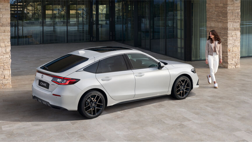 Lady standing next to rear three quarters view of Honda Civic e:HEV infront of building.