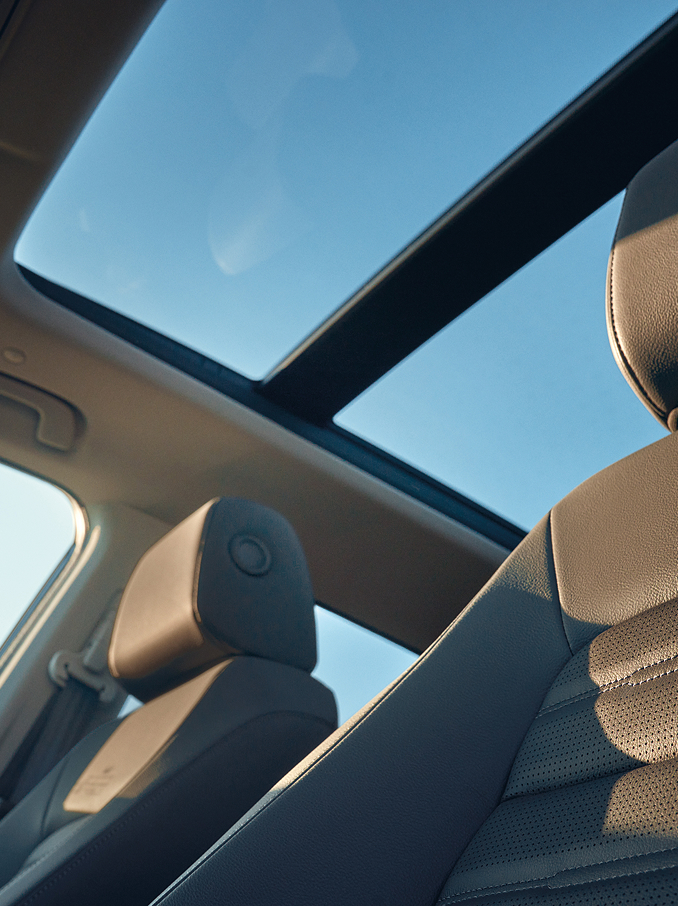 Interior view of a car showing a panoramic glass roof letting in blue sky and sunlight, with leather front seats and headrests illuminated by natural light.