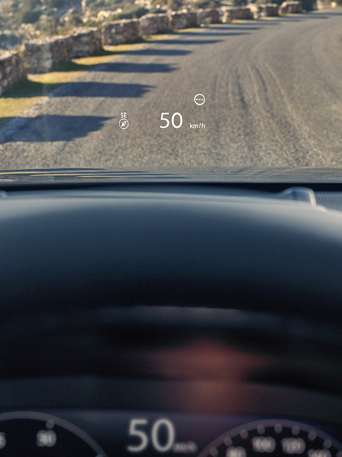 A first-person view from a driver's seat showing a Head-Up Display (HUD) projecting "50 km/h" and a compass onto the windshield above a gravel road.