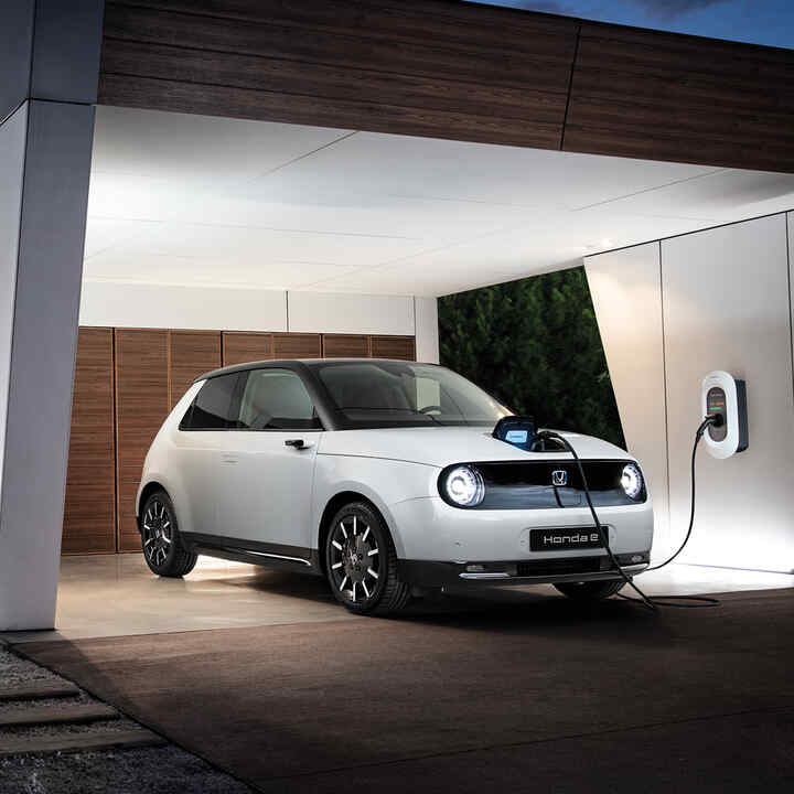 Front facing three quarter Honda e parked in the driveway charging as the owner enters the home.