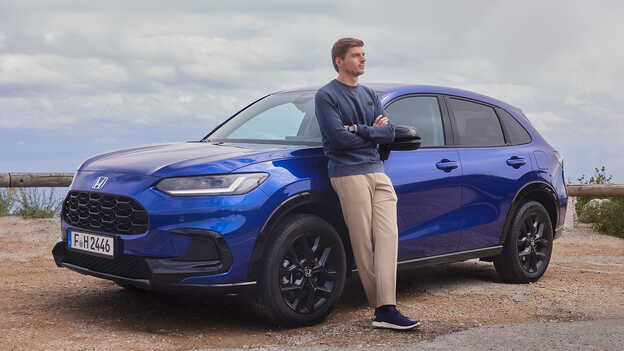 Max Verstappen leaning on blue Honda ZR-V Hybrid SUV car parked showcasing sporty aesthetic.