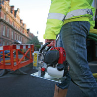 A man wearing blue jeans and yellow reflective jacket carries lightweight Honda water pump. 