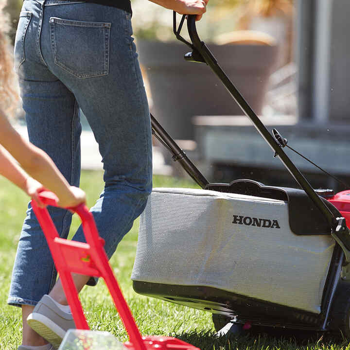 Child pushing toy lawnmower next to mother in garden location