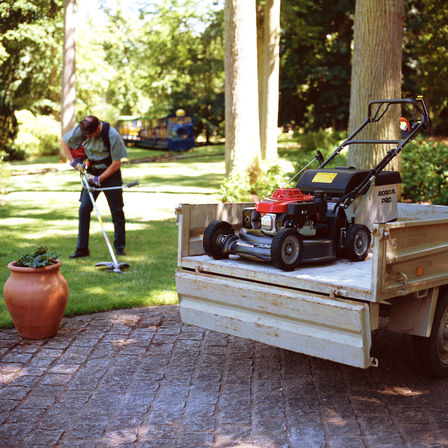 photo of honda hrh petrol lawnmower on trailer whilst man uses petrol strimmer on lawn in background