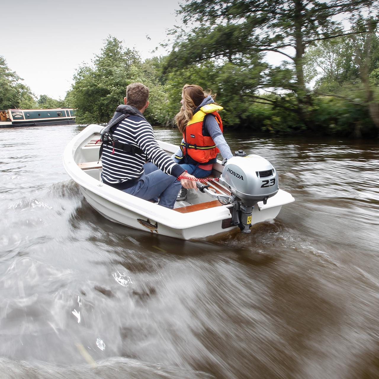 Two people in a boat using the Honda BF2.3 engine, next to a canal boat.