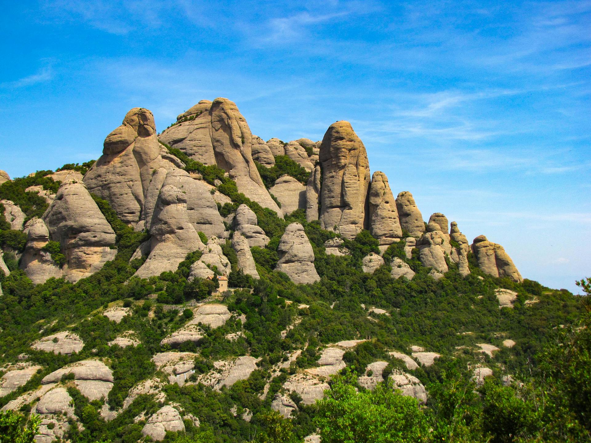 Rugged mountain landscape of Montserrat, Spain, featuring distinctive rounded rock formations and pinnacles rising above green vegetation against a blue sky.