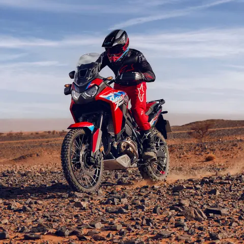 Model driving CRF1100L Africa twin motorbike on rocky terrain in desert location.