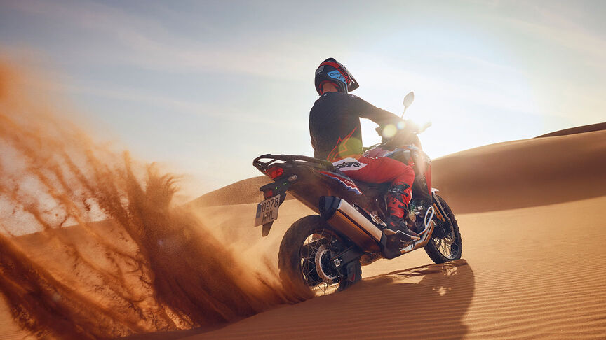 Model riding CRF1100L Africa Twin motorbike in a desert location.