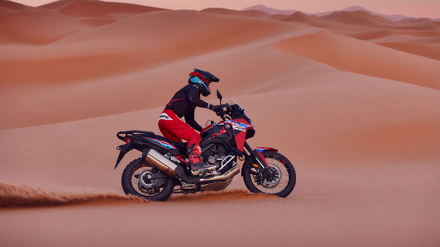 Model riding CRF1100L Africa Twin motorbike in a desert location.