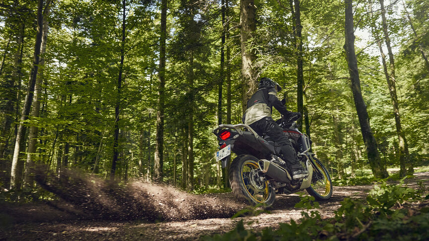 Model riding XL750 Transalp motorcycle in a forest location.