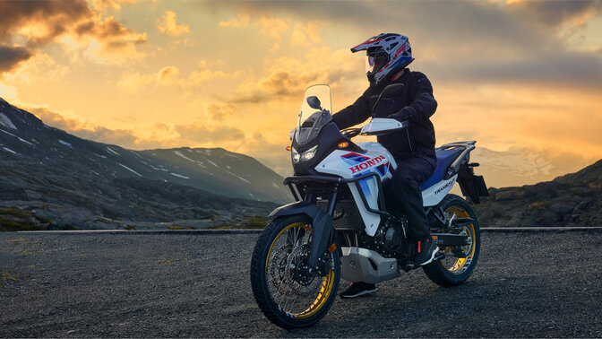 Model riding a XL750 Transalp motorcycle on the road in mountain location.
