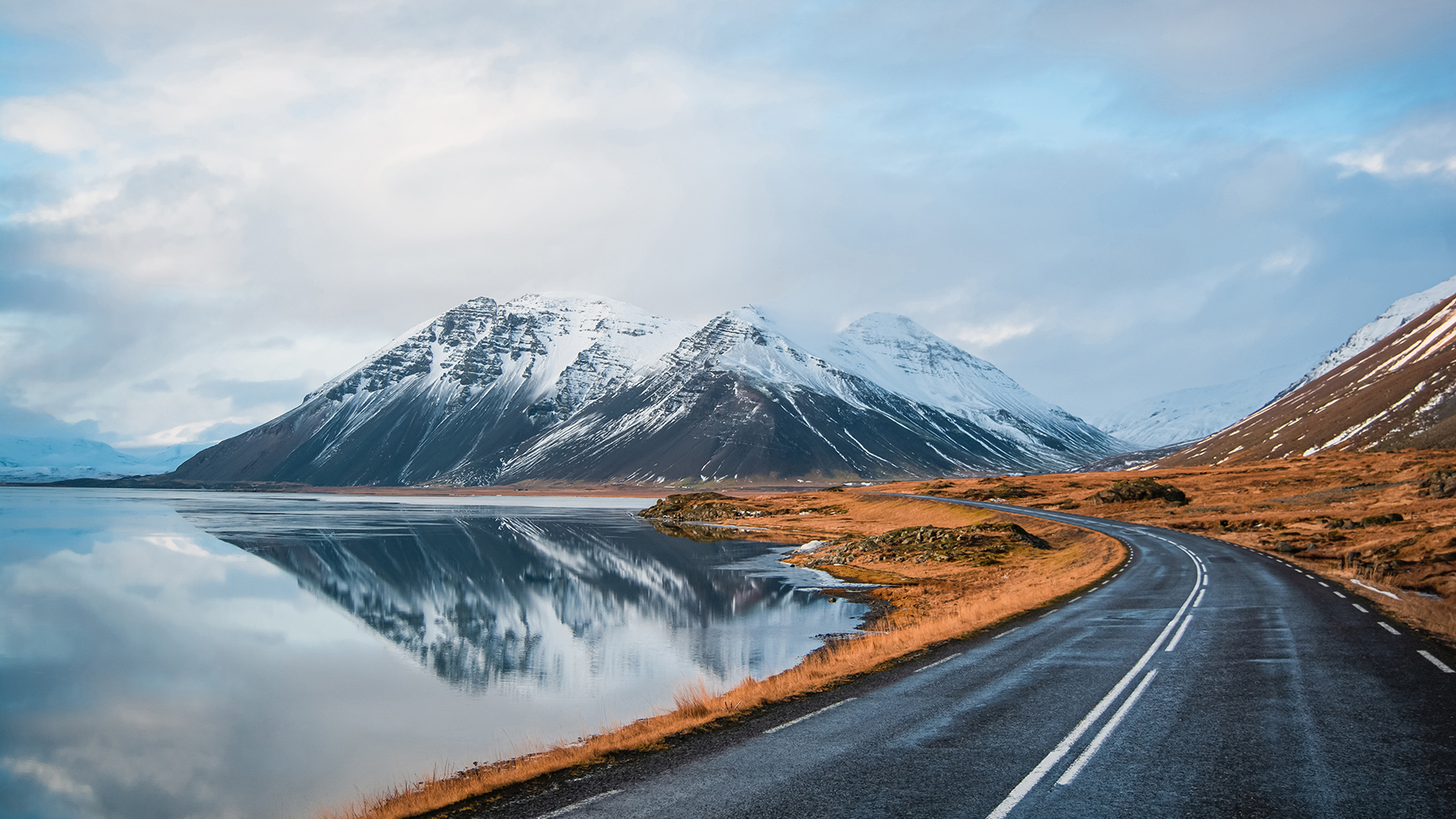Panoramic winter photo of road leading along coast of lake to volcanic mountains. High rocky peaks covered with snow layer mirroring on water surface. Driver's point of view on Ring road, Iceland.