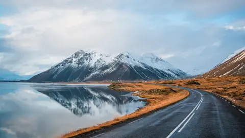 Panoramic winter photo of road leading along coast of lake to volcanic mountains. High rocky peaks covered with snow layer mirroring on water surface. Driver's point of view on Ring road, Iceland.