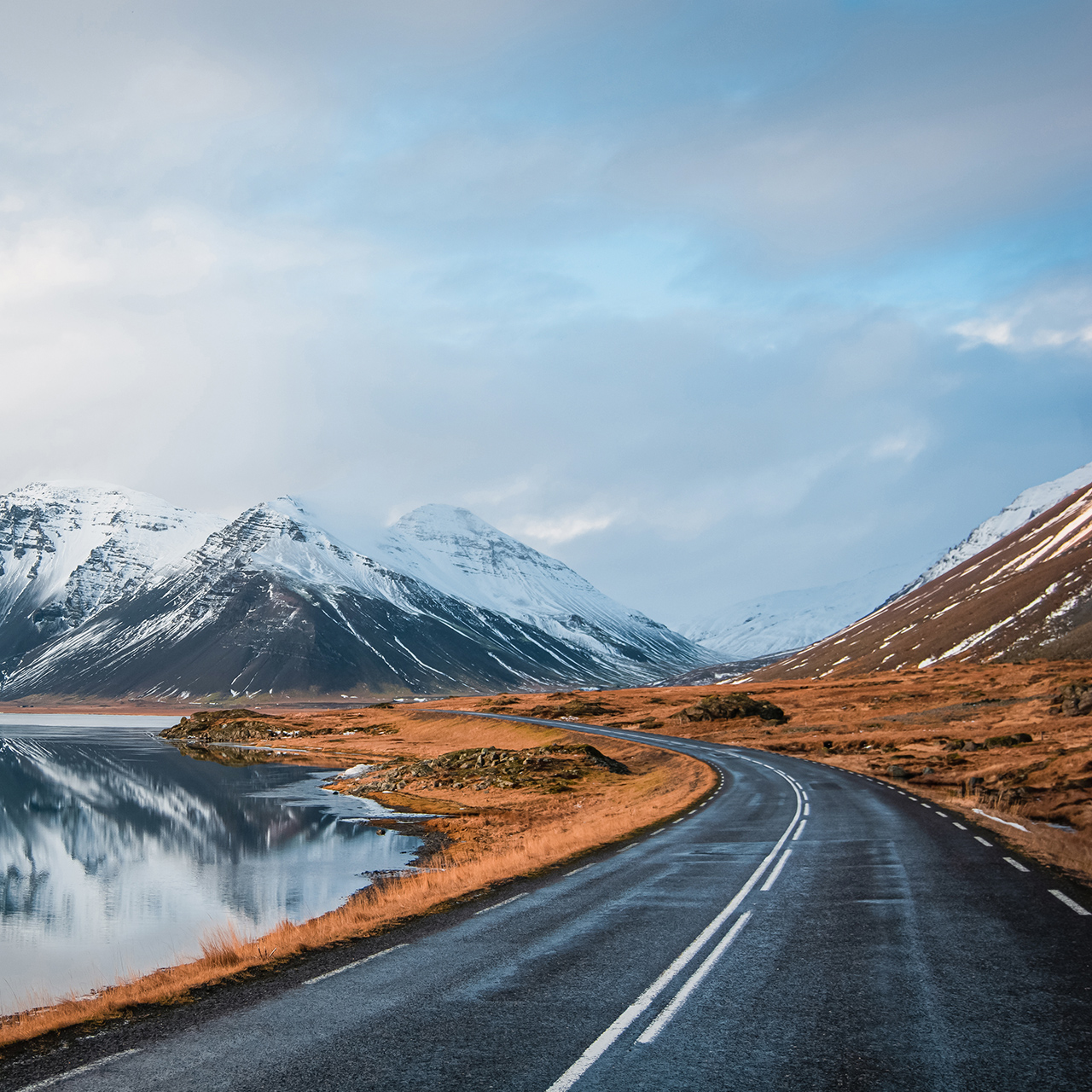 Panoramic winter photo of road leading along coast of lake to volcanic mountains. High rocky peaks covered with snow layer mirroring on water surface. Driver's point of view on Ring road, Iceland.