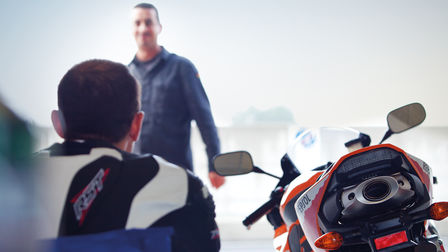 Rear of motorcycle, rider sitting on chair beside it. Man standing opposite the rider.