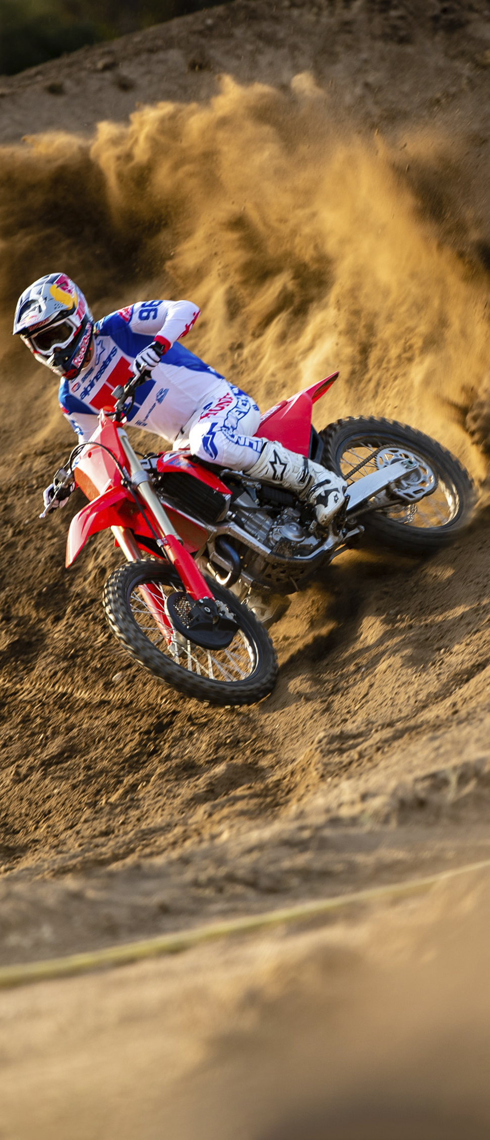 A motocross rider in white, blue, and red gear leans a red dirt bike into a steep sandy berm, spraying a large wave of dirt into the air as they power through the turn.