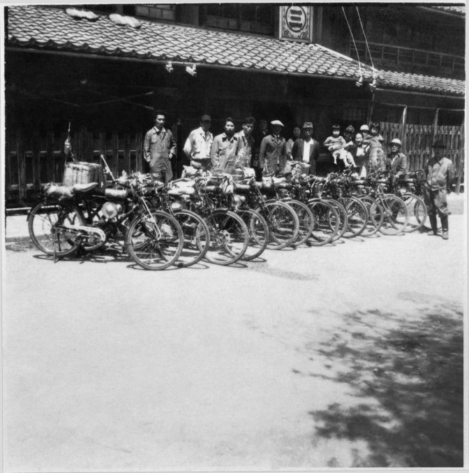 A row of 13 early Honda Motorcycles in Japan with people standing behind them