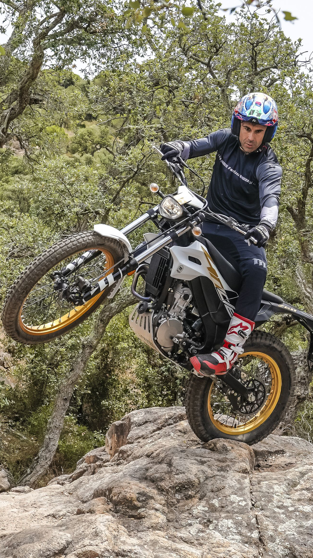 Rider lifting the front wheel of a Montesa 4RIDE motorcycle on a rocky forest path.