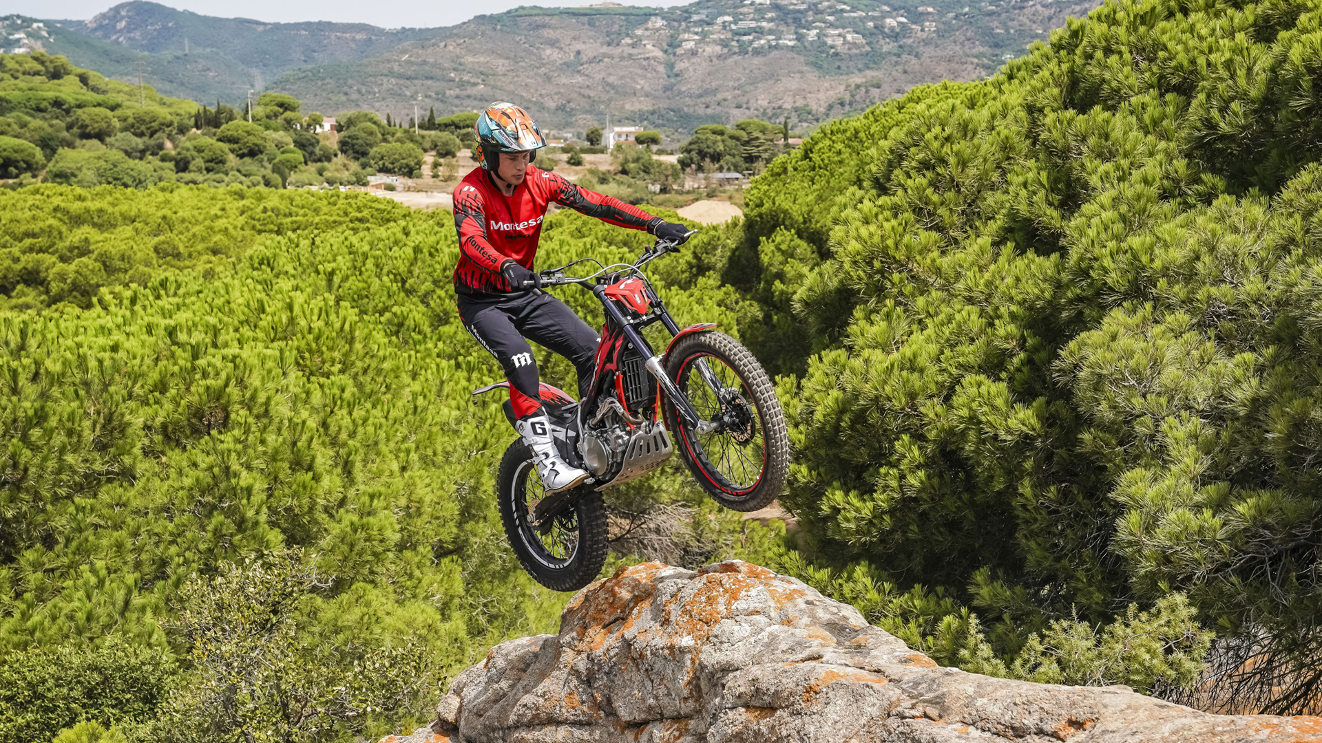 Rider jumping a red Montesa Cota 4RT 260R motorcycle off a rocky ledge over a scenic pine forest landscape.