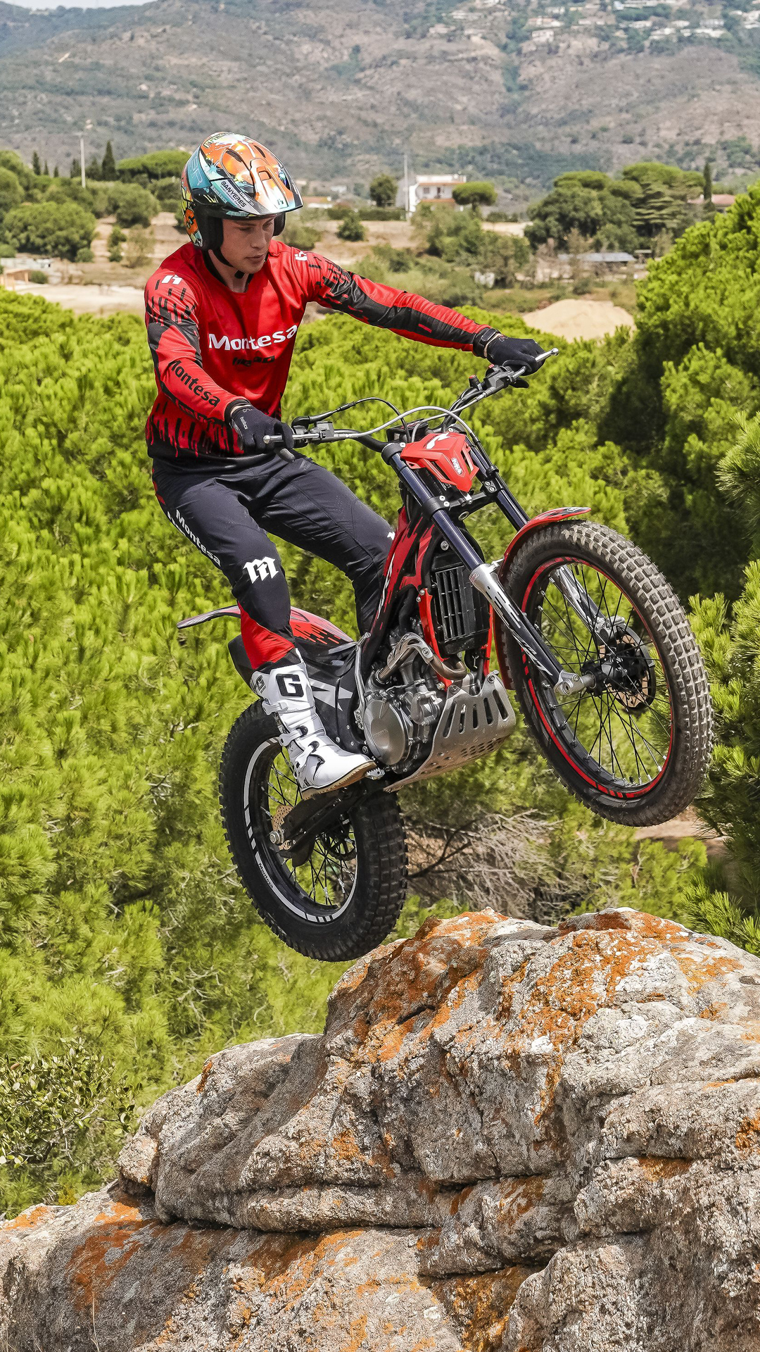Rider jumping a red Montesa Cota 4RT 260R motorcycle off a rocky ledge over a scenic pine forest landscape.