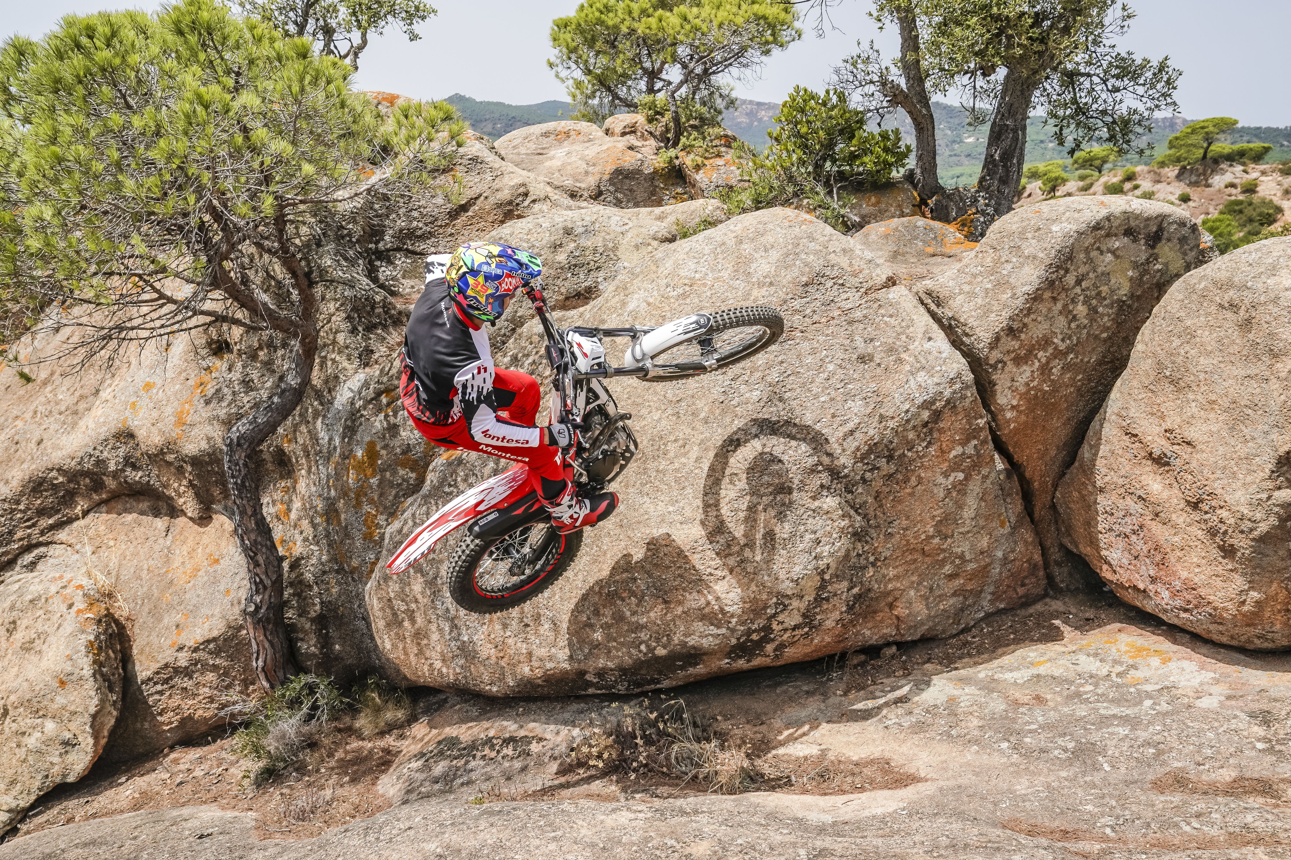Motorcycle rider performing a wheelie on a Montesa Cota 301RR bike on a rock formation.