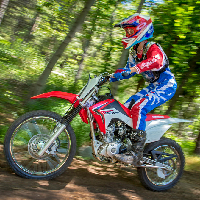 Side on view of CRF125F with rider on dirt track in forest race location. Left facing.