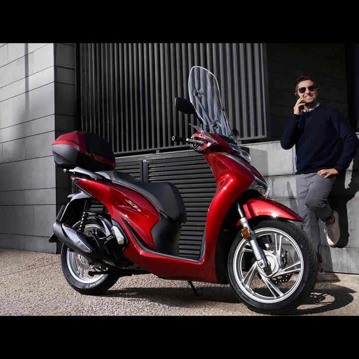 Man on phone stood next to Honda scooter.