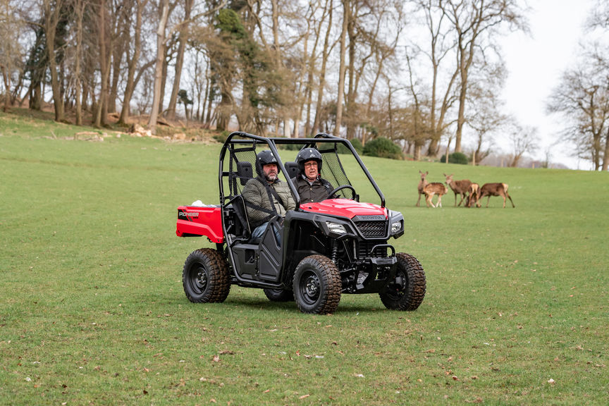 Picture of 2 people sat in a Honda Pioneer 520 with side by side seating, on a grass landscape with deer in the background