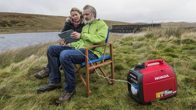 two people sitting on camp chairs using honda portable generator to power a tablet outdoors