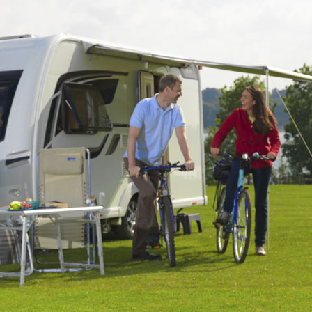two people on bicycles on a campsite with a white caravan and camp chair in the background