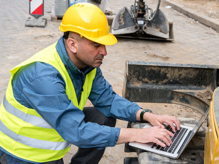 a worker wearing yellow safety vest and helmet using laptop on a building site