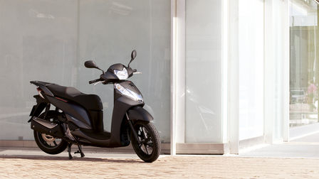 Rear of motorcycle, rider sitting on chair beside it. Man standing opposite the rider.